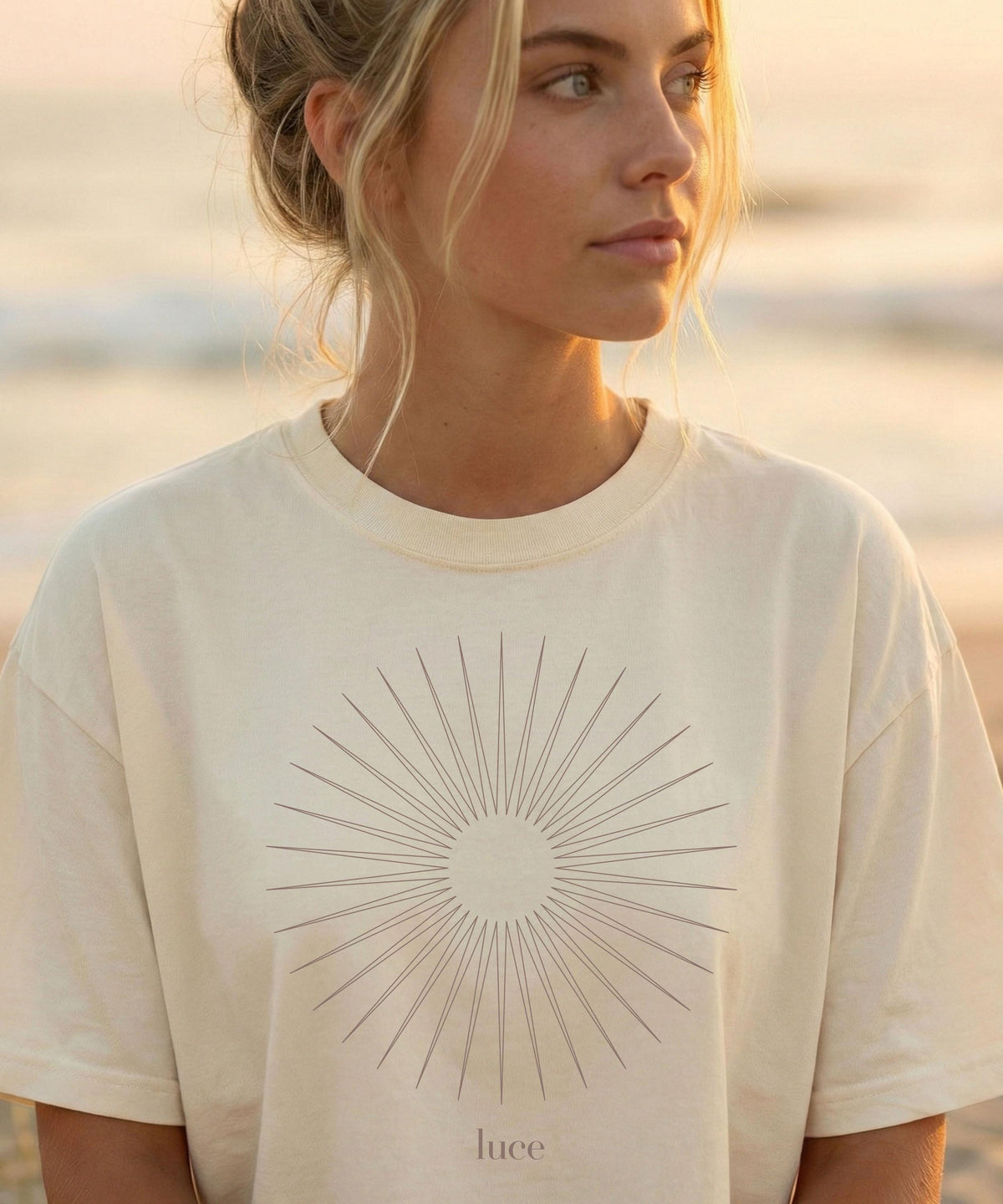 May include: Cream-colored t-shirt featuring a sunburst design with radiating lines and the word "luce" in a minimalist font. The shirt has a classic crew neck and short sleeves. The overall aesthetic is simple and elegant.