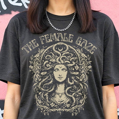 May include: Dark gray t-shirt featuring the text "THE FEMALE GAZE" above a detailed illustration of Medusa. The artwork includes a crescent moon and floral elements, creating a vintage, artistic aesthetic. The shirt is a casual wear item.