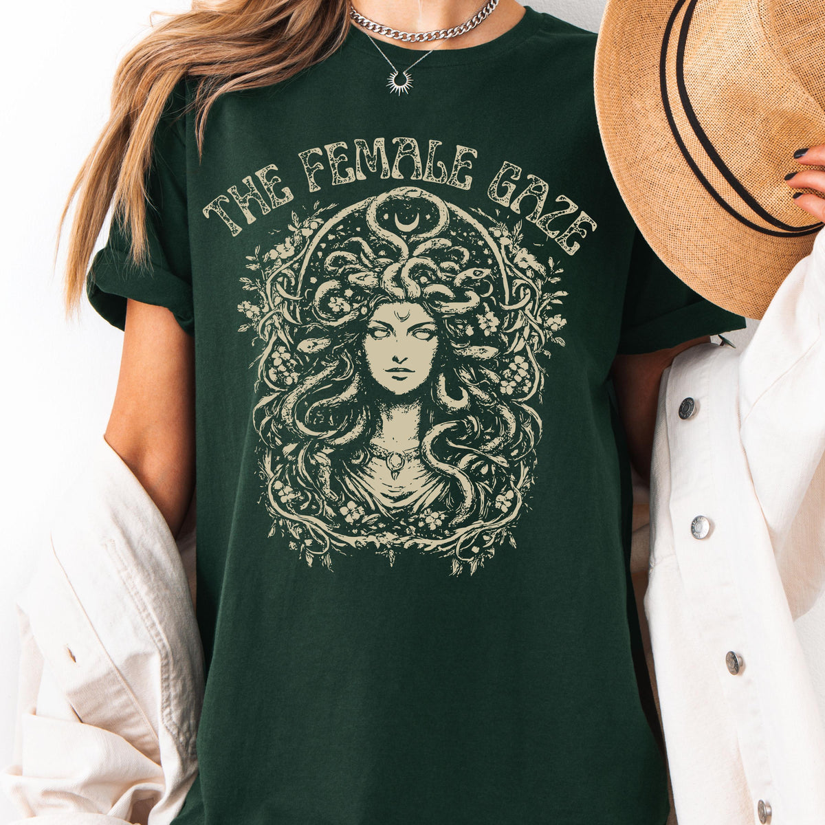 May include: Dark green t-shirt with the text "THE FEMALE GAZE" above a detailed illustration of Medusa. The illustration is in a cream color and features Medusa's face surrounded by snakes and floral elements.