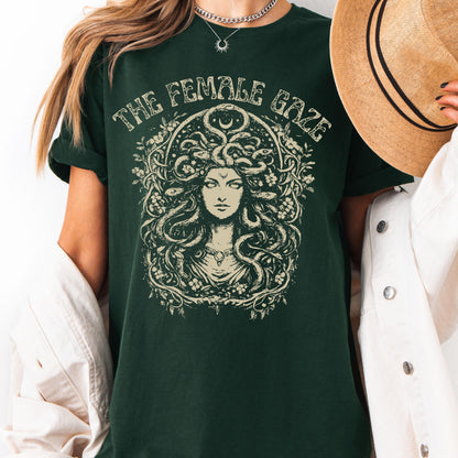 May include: Dark green t-shirt with the text "THE FEMALE GAZE" above a detailed illustration of Medusa. The illustration is in a cream color and features Medusa's face surrounded by snakes and floral elements.