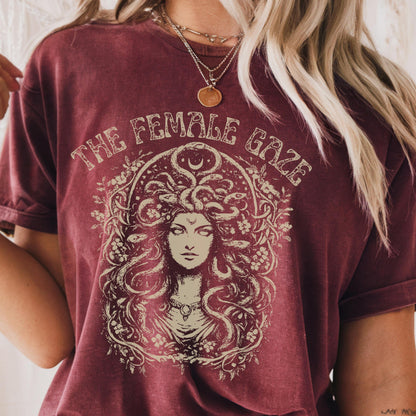 May include: Burgundy t-shirt featuring a detailed illustration of Medusa with the text "THE FEMALE GAZE" in a vintage style. The design is centered on the front of the shirt. The shirt has short sleeves and a relaxed fit.