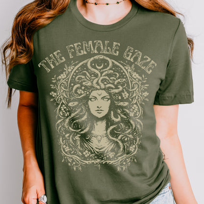 May include: Olive green t-shirt featuring a detailed illustration of a woman with snakes for hair, surrounded by floral elements. The text "THE FEMALE GAZE" is arched above the illustration in a vintage style font. The shirt has short sleeves.