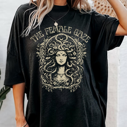 May include: Black t-shirt featuring a cream-colored graphic of Medusa with the text "THE FEMALE GAZE" in an arched design above the image. The graphic is surrounded by floral and crescent moon elements.