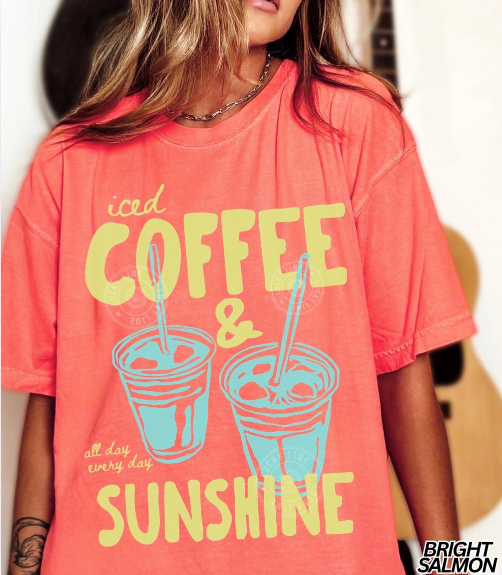 May include: A coral-colored t-shirt with the words "iced COFFEE & SUNSHINE" in yellow and a graphic of two iced coffee drinks in blue. The phrase "all day every day" is printed below the drinks.
