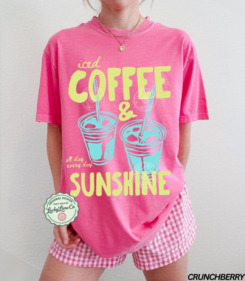 May include: A pink t-shirt with the words "iced COFFEE & SUNSHINE" in yellow and teal. The shirt also features two iced coffee cup illustrations. The words "all day every day" are printed below the cups. The shirt is paired with pink and white checkered shorts.
