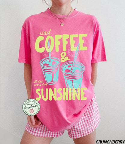 May include: A pink t-shirt with the words "iced COFFEE & SUNSHINE" in yellow and teal. The shirt also features two iced coffee cup illustrations. The words "all day every day" are printed below the cups. The shirt is paired with pink and white checkered shorts.