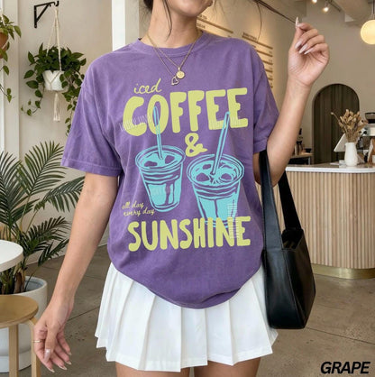 May include: A purple t-shirt with the words "iced COFFEE & SUNSHINE" in yellow and teal. The shirt features a graphic of two iced coffee drinks. The text "all day every day" is below the graphic. The shirt is paired with a white pleated skirt.