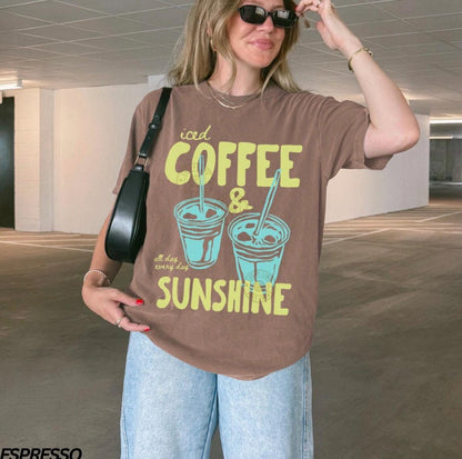 May include: Brown t-shirt with the words "iced COFFEE & SUNSHINE" in yellow and two turquoise iced coffee cup graphics. The shirt also has the words "all day every day" in a smaller font. The person is wearing blue jeans and sunglasses.