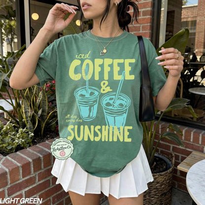 May include: Light green t-shirt with the text "iced COFFEE & SUNSHINE" in yellow and teal. The shirt features two iced coffee cup illustrations. The shirt is paired with a white pleated skirt.