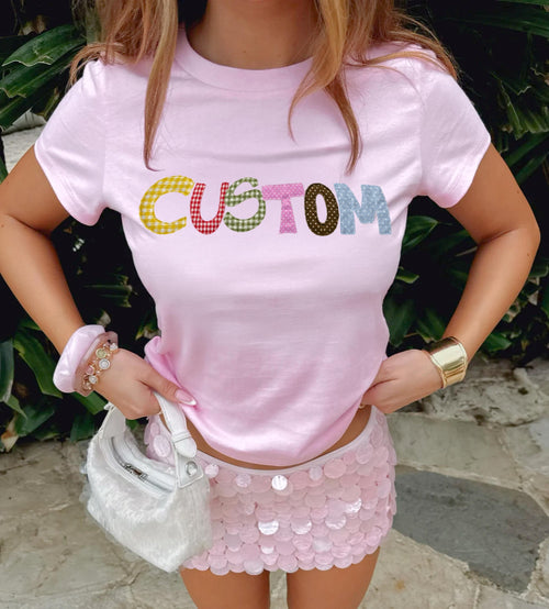 May include: A light pink t-shirt with the word "CUSTOM" in colorful printed patchwork letters. The model is wearing a matching pink sequin mini skirt, a white handbag, and gold jewelry.