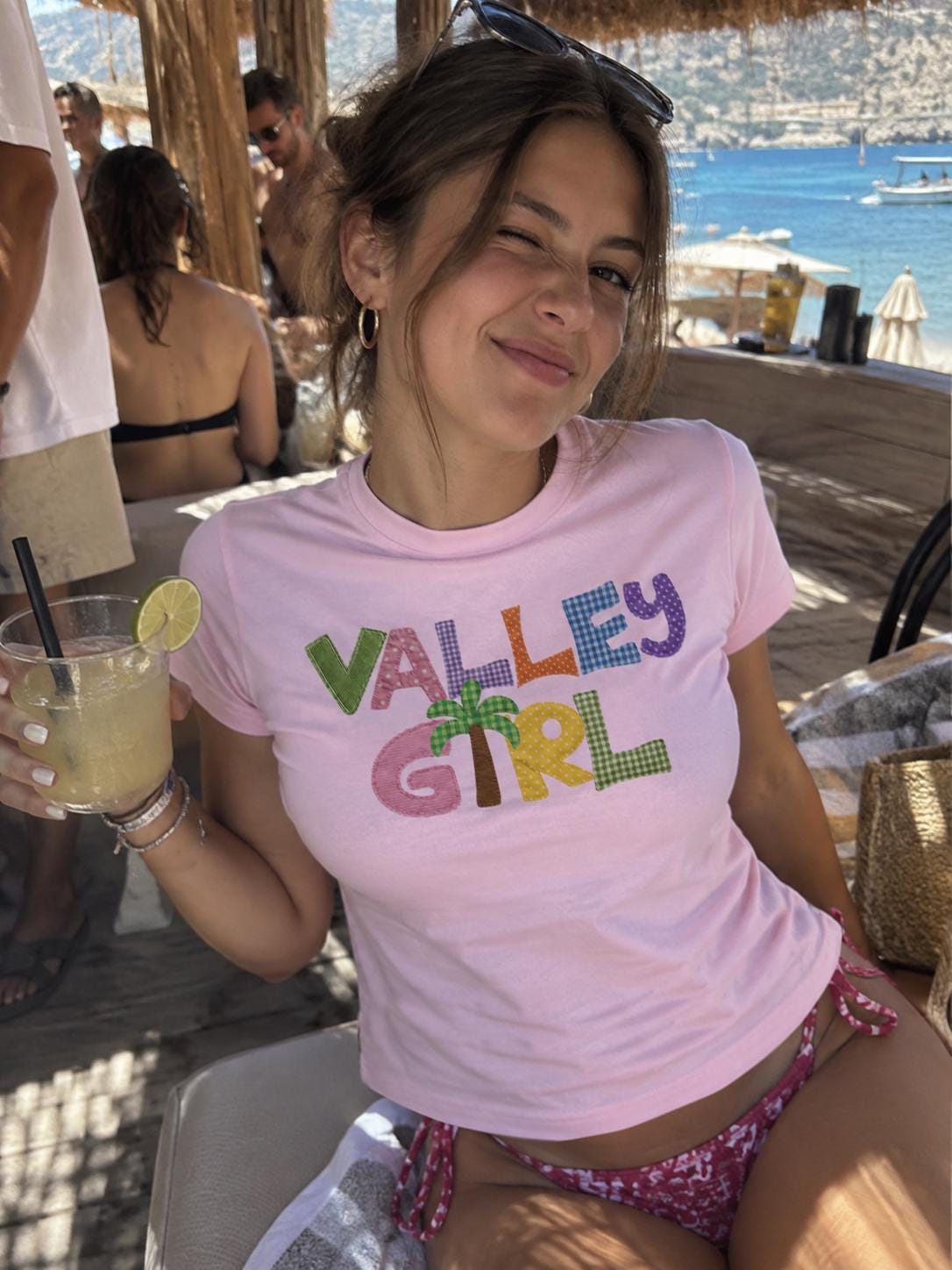 May include: A light pink short-sleeved t-shirt featuring the text VALLEY GIRL in colorful, patterned fabric letters. The letter I is replaced by a brown and green palm tree graphic. The shirt has a crew neckline and a fitted silhouette.