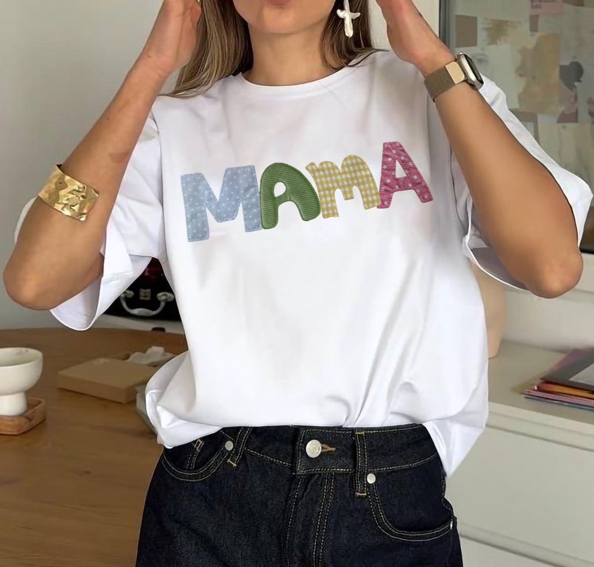 May include: White t-shirt with the word "MAMA" in colorful, patchwork-style printed letters. The letters are in blue with white polka dots, green gingham, yellow gingham, and pink floral patterns. The shirt is paired with dark wash denim jeans.