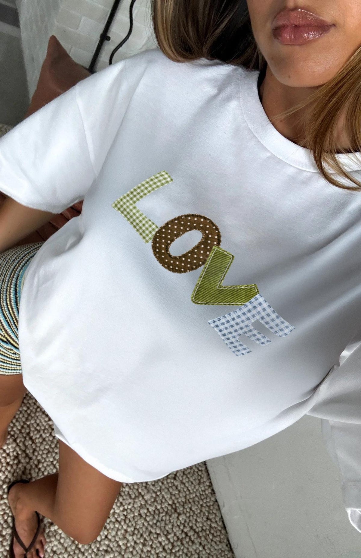May include: White t-shirt with the word "LOVE" patchwork letters printed on the front. The letters are in various patterns: green gingham, brown polka dots, green ribbon, and blue and white gingham. The shirt is a casual style.
