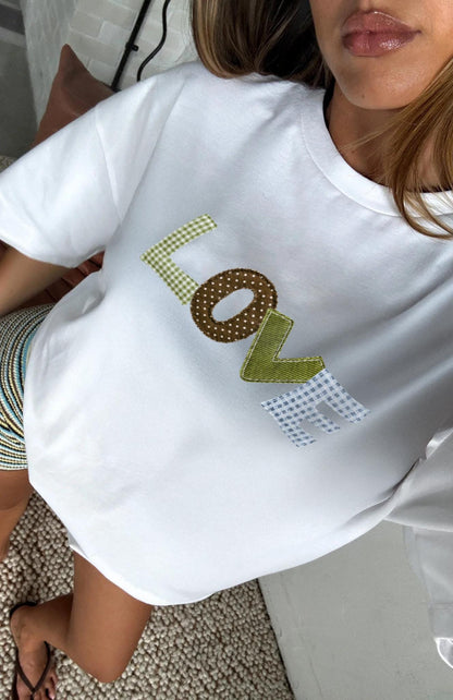 May include: White t-shirt with the word "LOVE" patchwork letters printed on the front. The letters are in various patterns: green gingham, brown polka dots, green ribbon, and blue and white gingham. The shirt is a casual style.