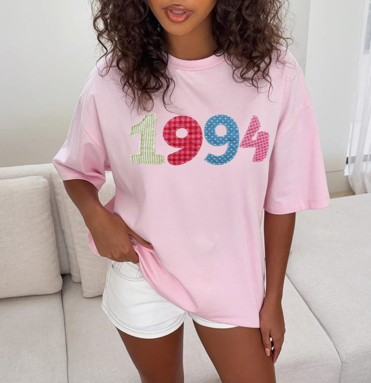 May include: A light pink t-shirt with the year "1994" in colorful printed patchwork lettering. The numbers are in green, red gingham, blue polka dots, and pink floral patterns. The shirt has short sleeves and a relaxed fit.