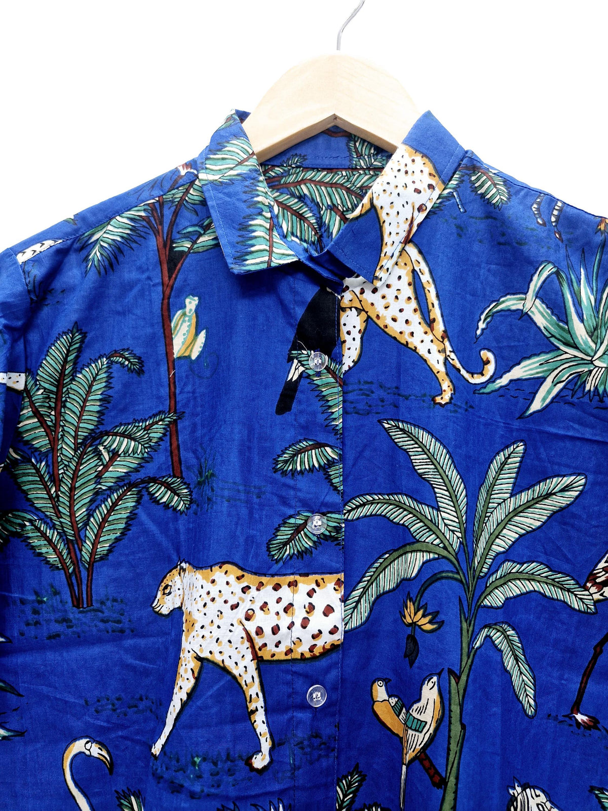 May include: A blue button-down shirt with a tropical print featuring leopards, palm trees, and birds. The shirt is hanging on a wooden hanger. The fabric has a crinkled texture.