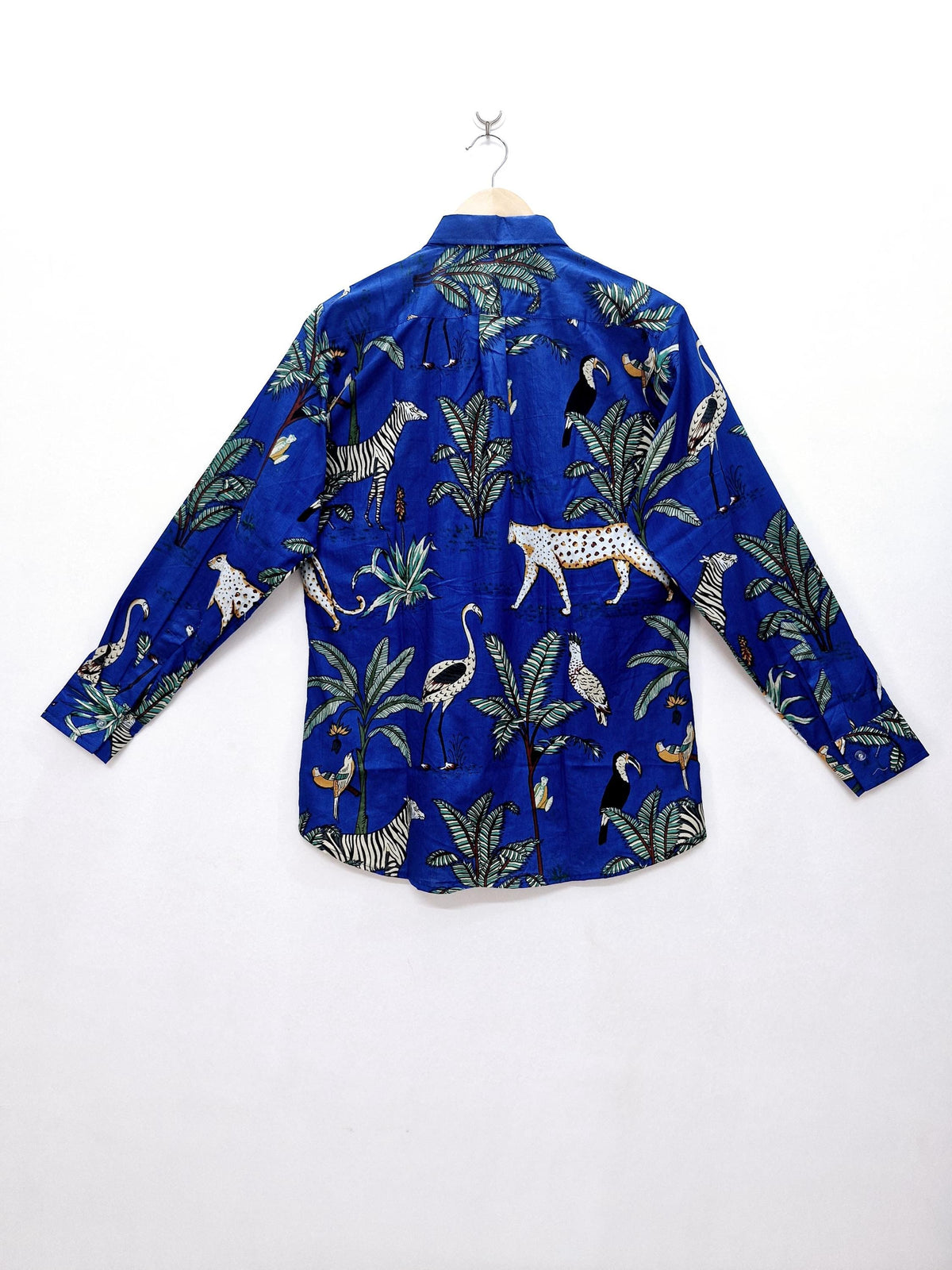 May include: A long-sleeved blue button-down shirt with a vibrant jungle-themed print. The design features zebras, leopards, toucans, and other tropical elements amidst palm trees. The shirt is hanging on a wooden hanger.