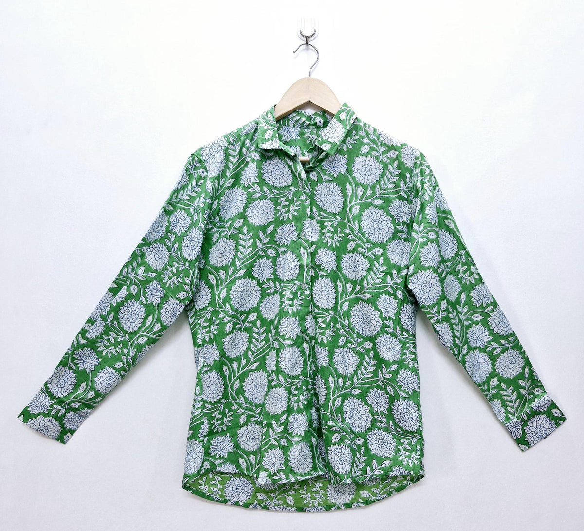 May include: A long-sleeved button-down shirt with a green background and a white floral pattern. The shirt is hanging on a wooden hanger against a white background. The shirt's design features a repeating pattern of flowers and leaves.