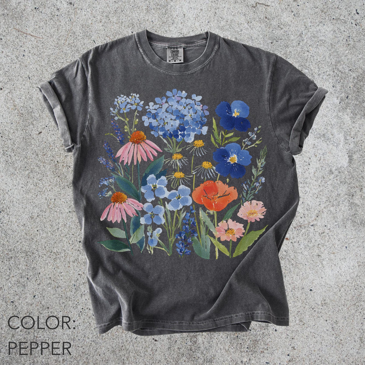 May include: A dark gray t-shirt featuring a watercolor floral design. The design includes pink coneflowers, blue hydrangeas, orange poppies, and other colorful flowers. The shirt's color is described as "pepper".