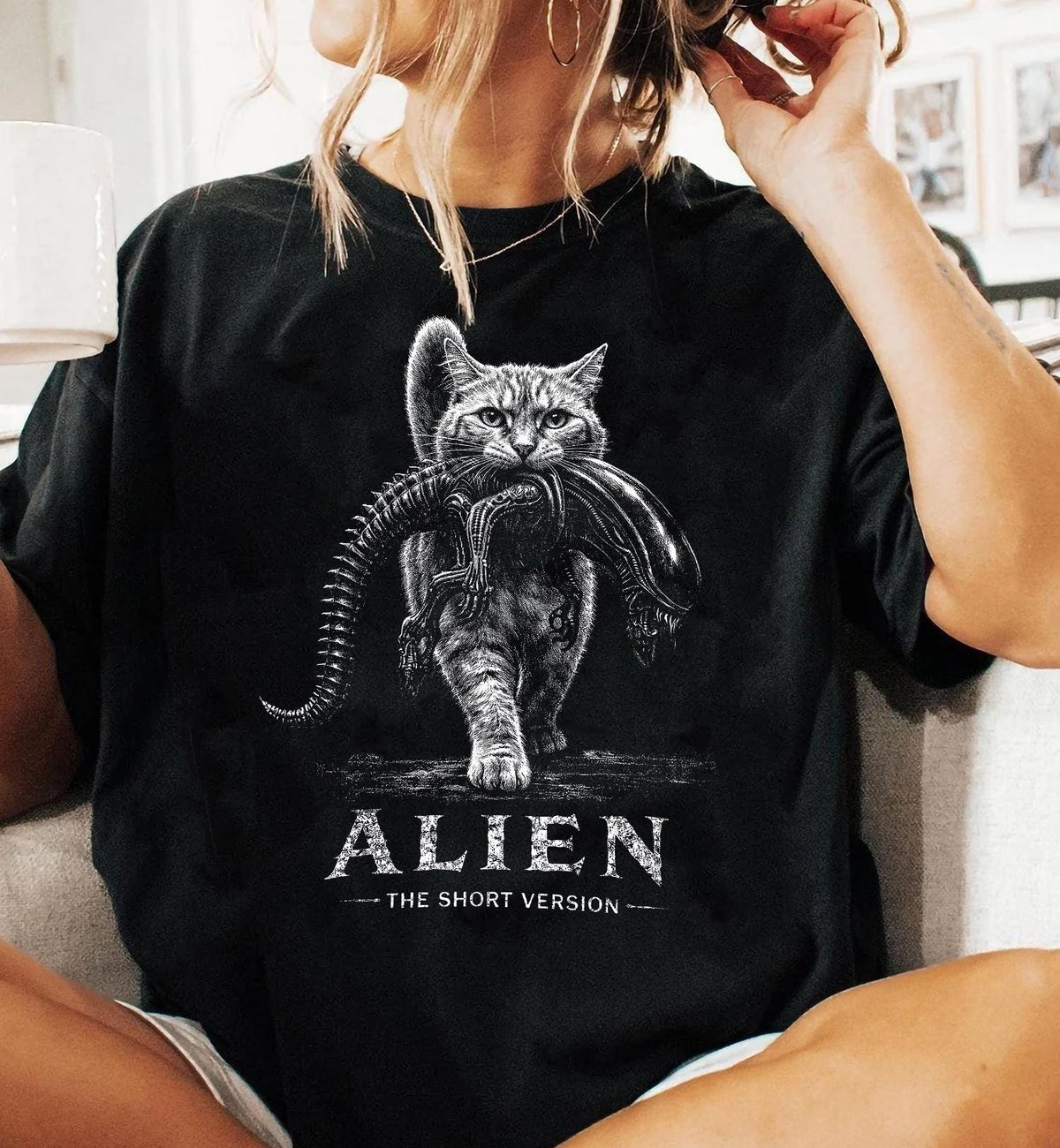 May include: A black t-shirt featuring a white graphic illustration of a tabby cat carrying a small xenomorph creature in its mouth. Below the cat, the text reads ALIEN THE SHORT VERSION in a distressed white font, centered on the chest area.