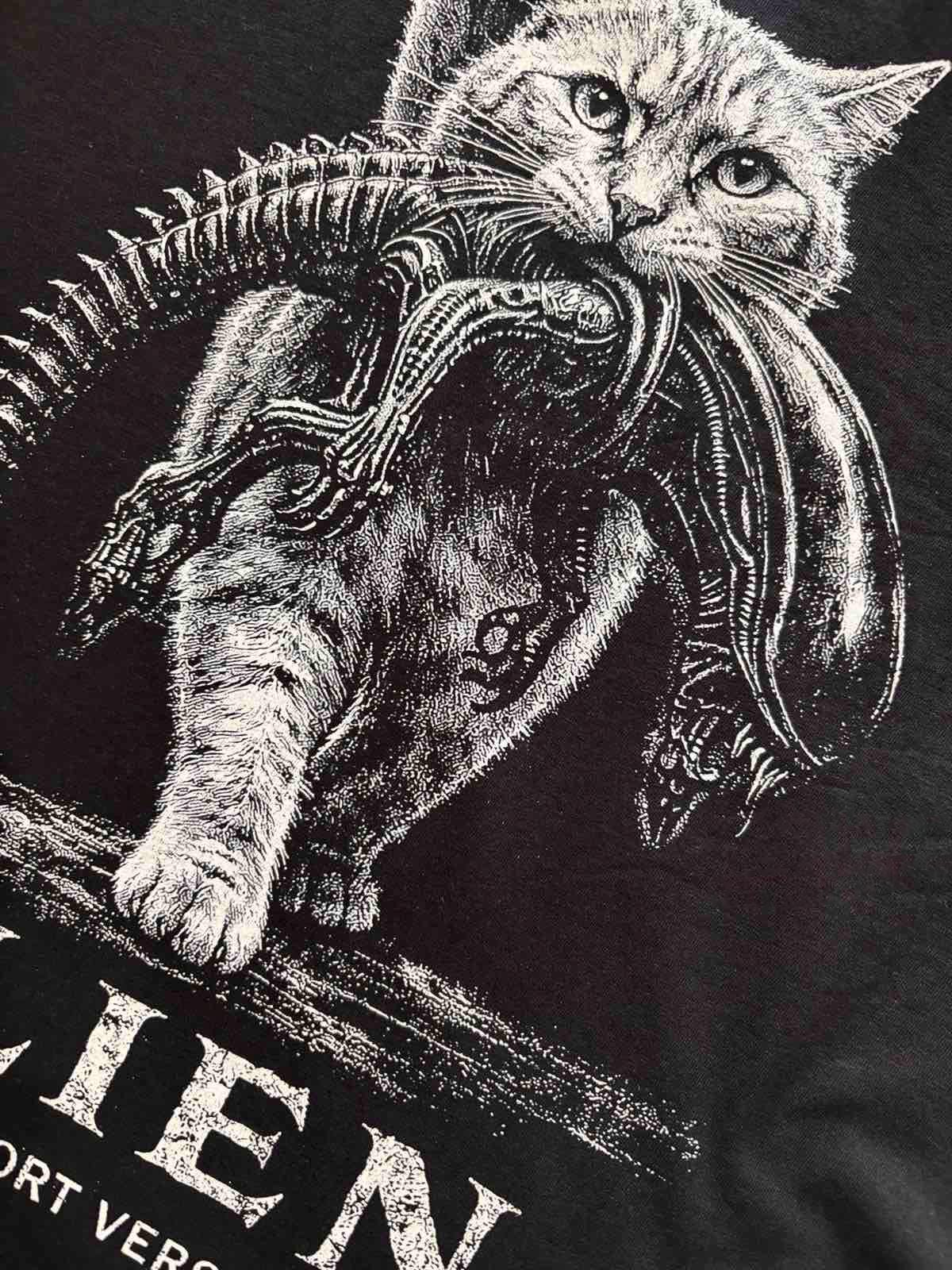 May include: A black t-shirt featuring a white graphic illustration of a cat carrying a xenomorph creature in its mouth. The design includes the partial text "IEN" and "ORT VER" in a distressed white font at the bottom of the printed graphic.