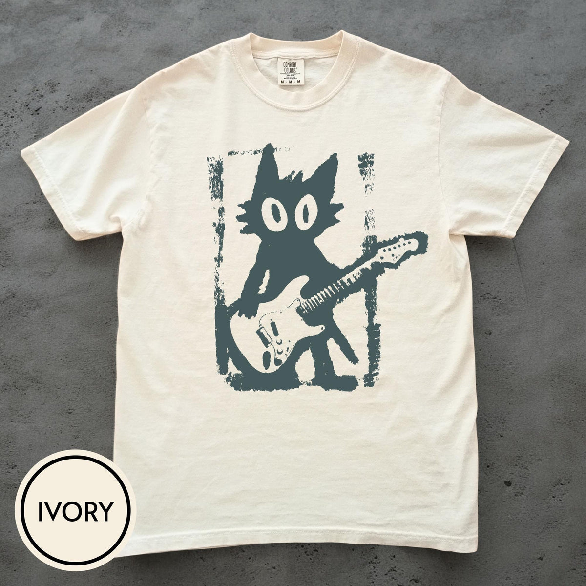 May include: An ivory short-sleeve t-shirt featuring a dark gray graphic print of a stylized cat playing an electric guitar. The cat is centered within a rectangular, distressed-edge frame. The inner neck label displays the text Comfort Colors.