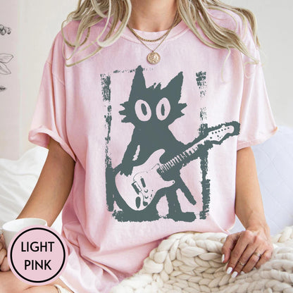 May include: A light pink short-sleeve t-shirt featuring a dark gray graphic of a stylized cat playing an electric guitar. The cat is depicted in a distressed, blocky style within a rectangular frame. The shirt has a crew neckline and a relaxed fit.