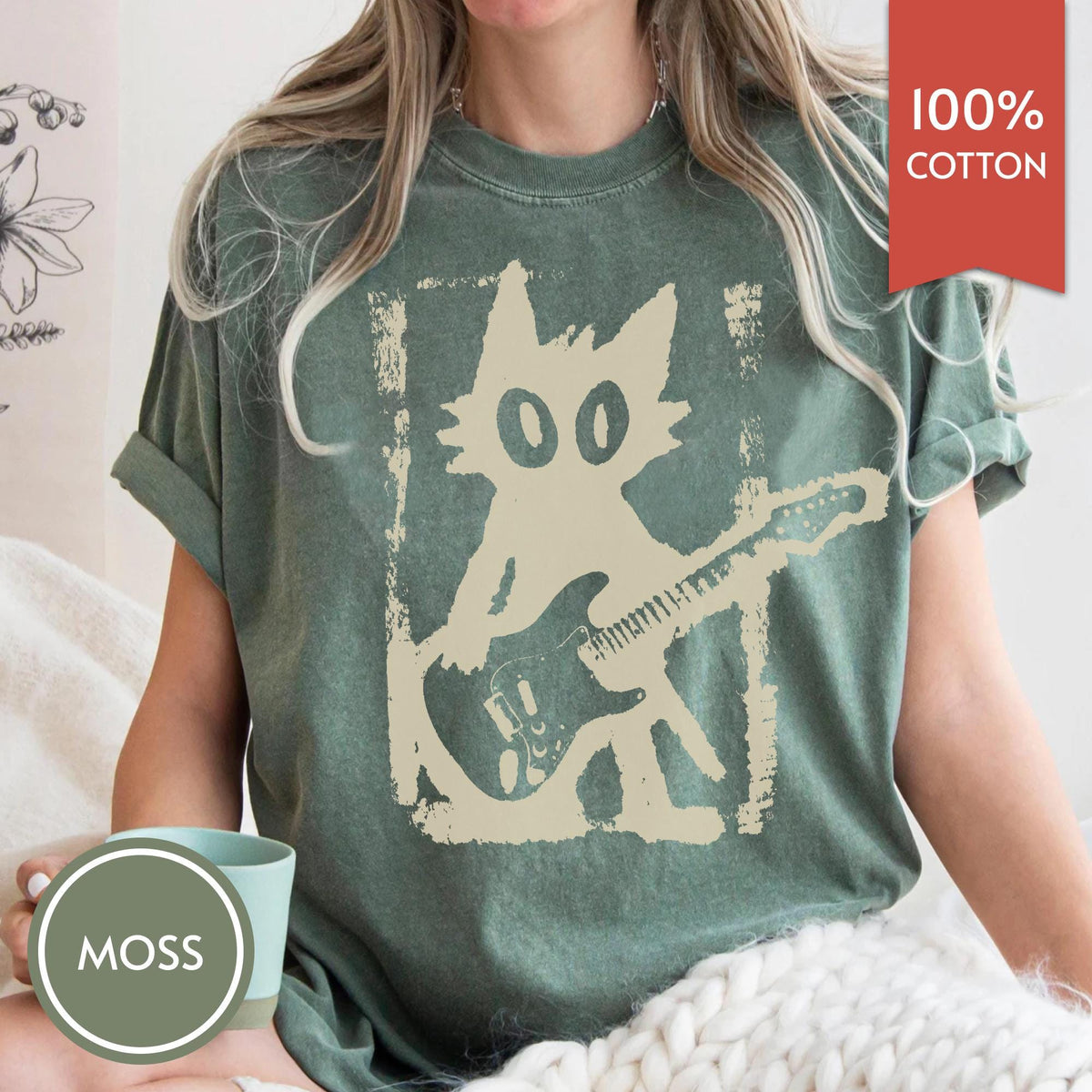 May include: A moss green t-shirt featuring a cream-colored graphic of a stylized cat playing an electric guitar. The design has a distressed, textured appearance. A red rectangular tag in the corner displays the text 100% COTTON.