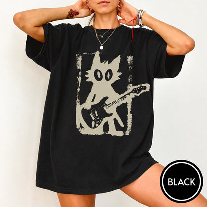 May include: A black short-sleeve t-shirt featuring a large, light-colored graphic print of a stylized cat playing an electric guitar. The cat illustration is framed within a rectangular, distressed-looking border, centered on the chest area.