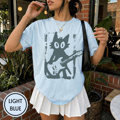 May include: A light blue t-shirt featuring a dark gray graphic of a cartoon cat playing an electric guitar. The cat has large oval eyes and is centered within a rectangular frame. The shirt has a crew neckline and short sleeves, paired with a white pleated skirt.