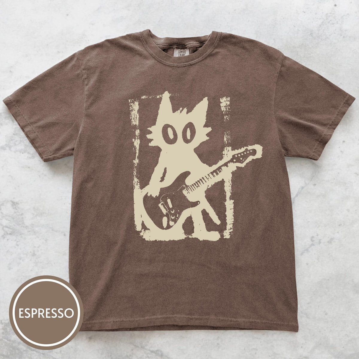 May include: A brown short-sleeve t-shirt featuring a cream-colored graphic of a cat playing an electric guitar inside a distressed rectangular frame. The shirt includes a circular badge in the bottom left corner with the text ESPRESSO.