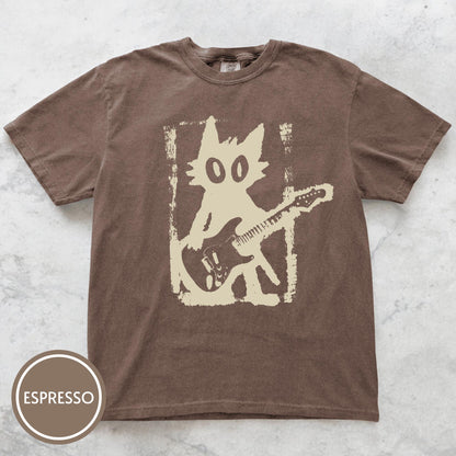 May include: A brown short-sleeve t-shirt featuring a cream-colored graphic of a cat playing an electric guitar inside a distressed rectangular frame. The shirt includes a circular badge in the bottom left corner with the text ESPRESSO.
