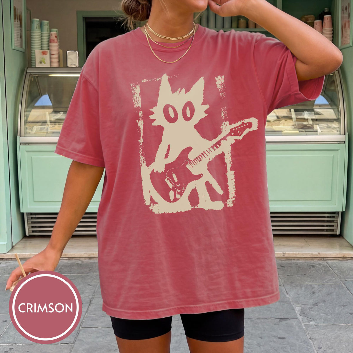 May include: A crimson short-sleeve t-shirt featuring a large, off-white graphic print of a stylized cat playing an electric guitar. The graphic has a distressed, textured appearance and is centered on the front of the garment. The shirt has a crew neckline.