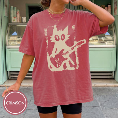 May include: A crimson short-sleeve t-shirt featuring a large, off-white graphic print of a stylized cat playing an electric guitar. The graphic has a distressed, textured appearance and is centered on the front of the garment. The shirt has a crew neckline.