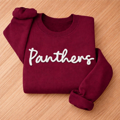 May include: A maroon sweatshirt with the word "Panthers" in white, cursive lettering. The sweatshirt is folded neatly, with the sleeves and collar visible. The fabric appears soft and comfortable.