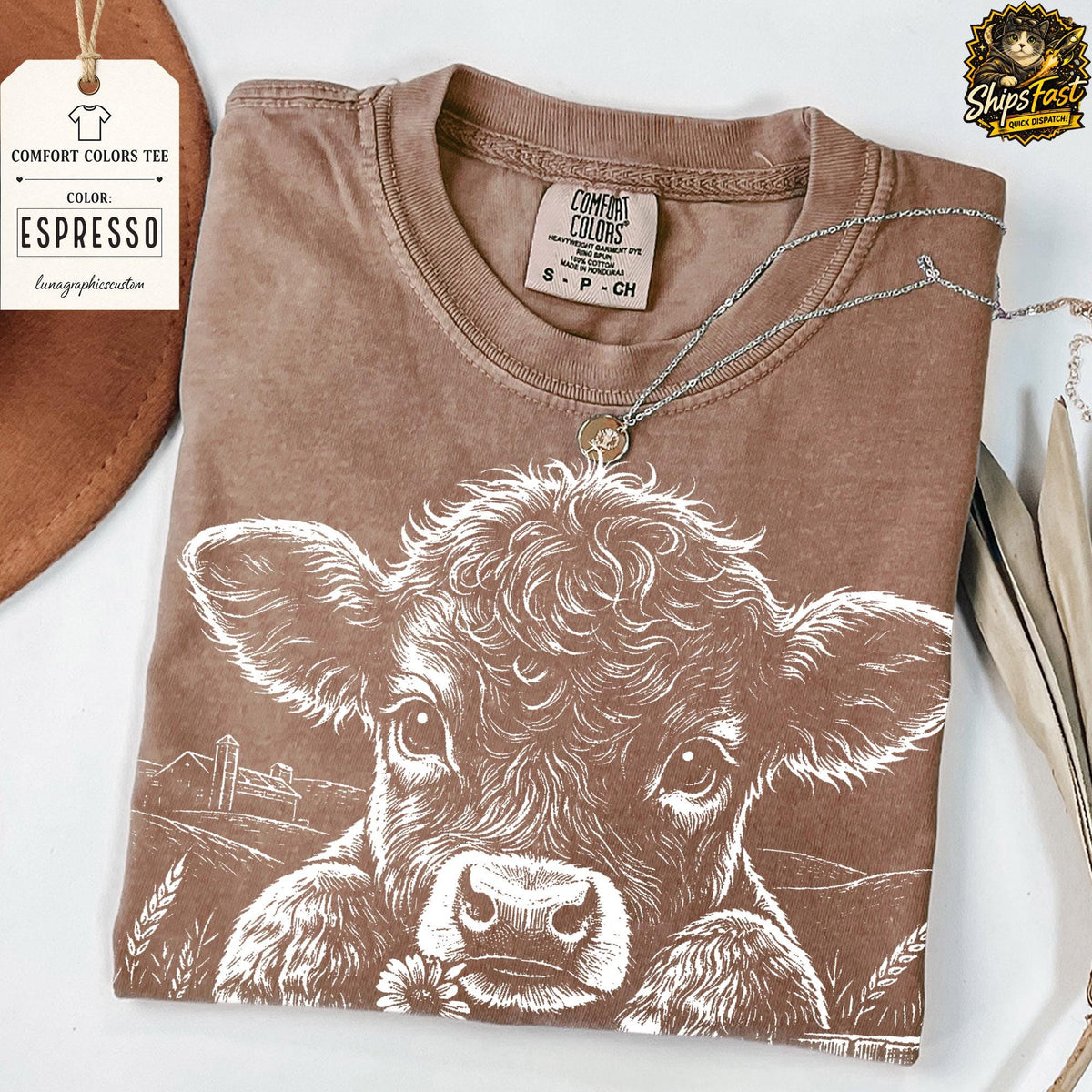 May include: Espresso-colored Comfort Colors tee featuring a white graphic of a cow with a flower. The shirt has a crew neck and short sleeves. A silver necklace with a pendant is draped across the shirt. A tag with the text "Comfort Colors Tee" is attached.