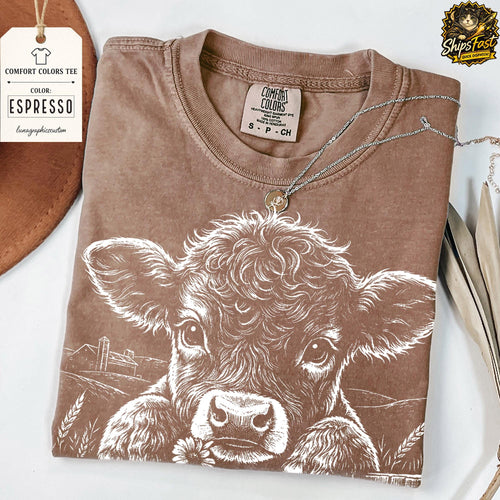 May include: Espresso-colored Comfort Colors tee featuring a white graphic of a cow with a flower. The shirt has a crew neck and short sleeves. A silver necklace with a pendant is draped across the shirt. A tag with the text "Comfort Colors Tee" is attached.