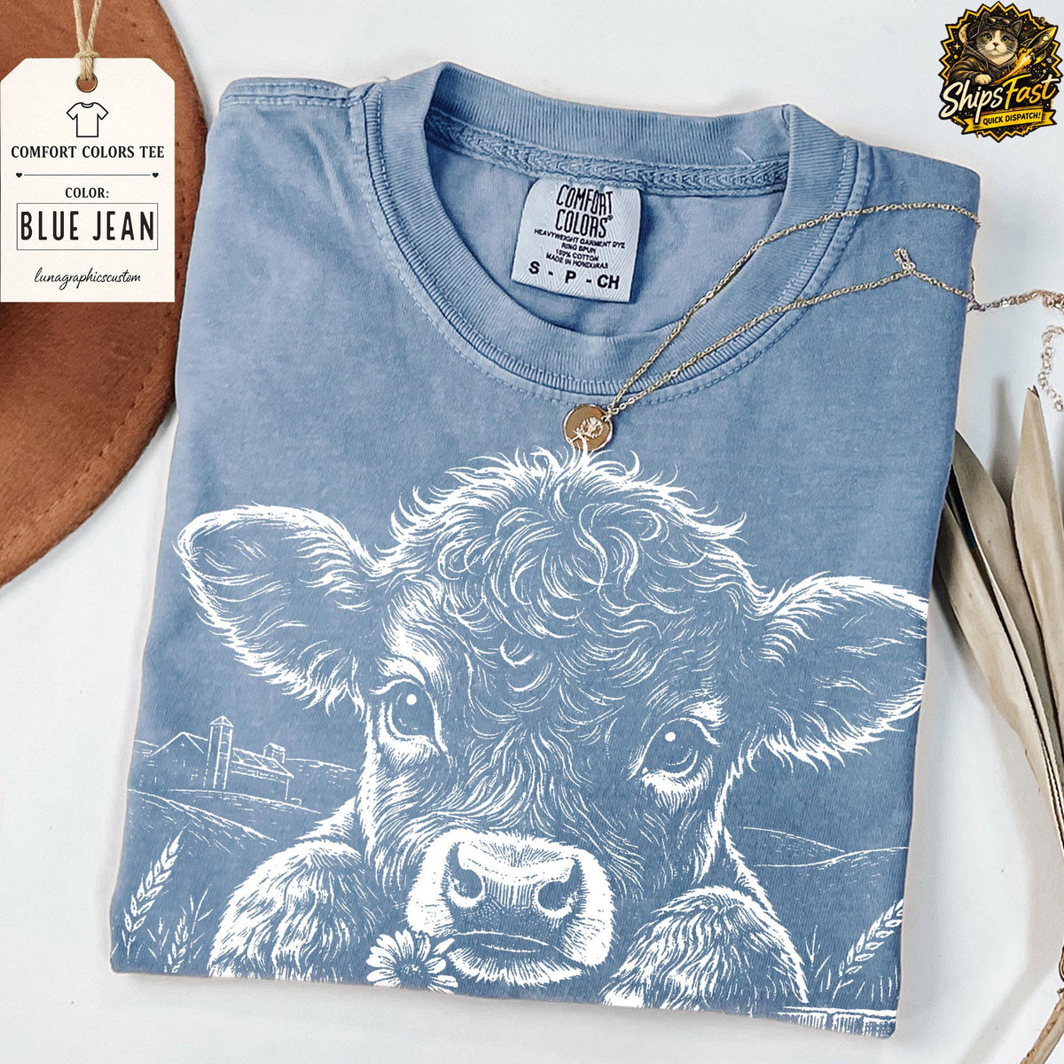May include: A blue jean Comfort Colors tee shirt with a white graphic of a cow's face. The cow is peeking over a field of wheat and holding a flower. A gold necklace is draped over the shirt.