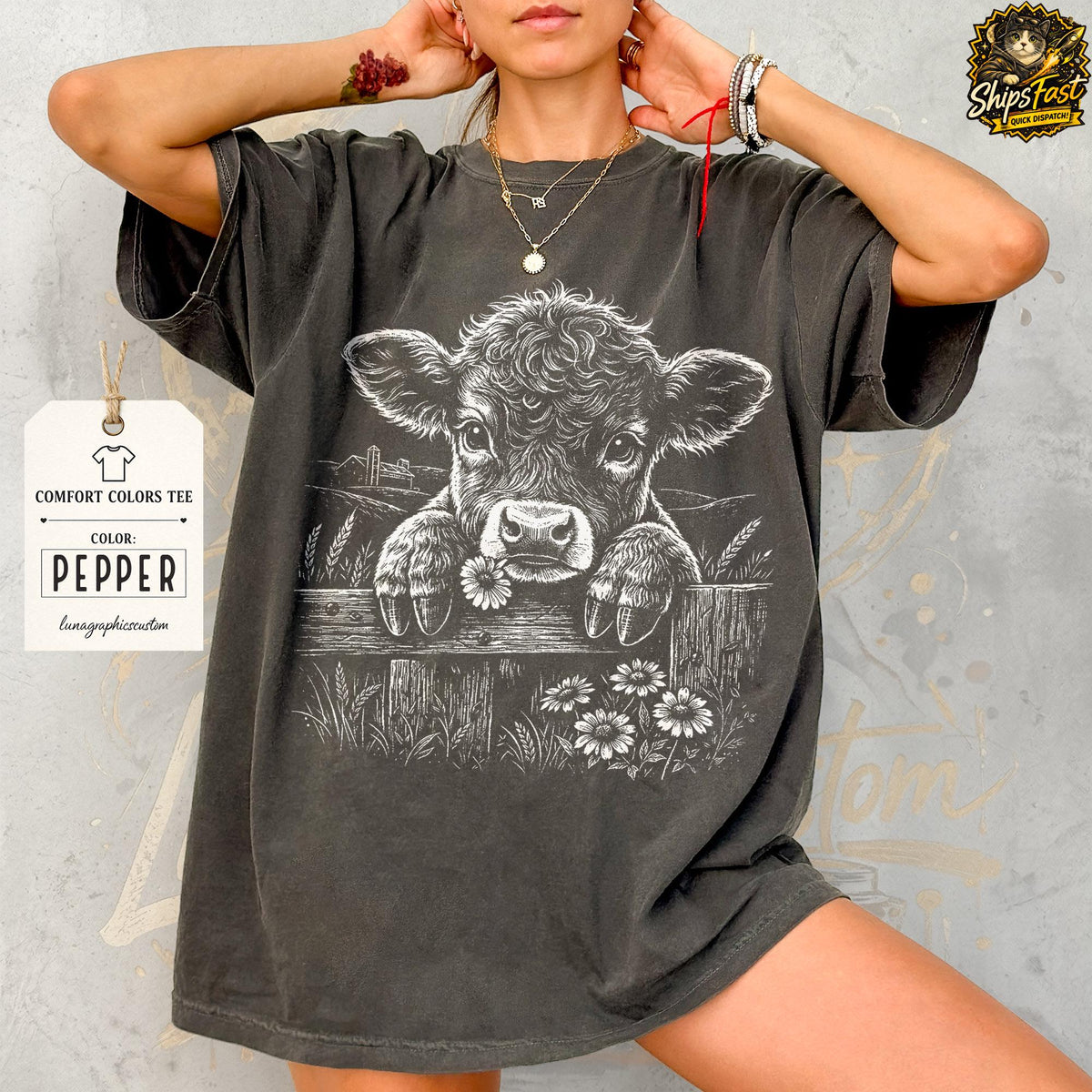 May include: Pepper-colored Comfort Colors tee featuring a detailed graphic of a cow peering over a wooden fence, surrounded by daisies and wheat. The design is in white, creating a striking contrast against the dark shirt.
