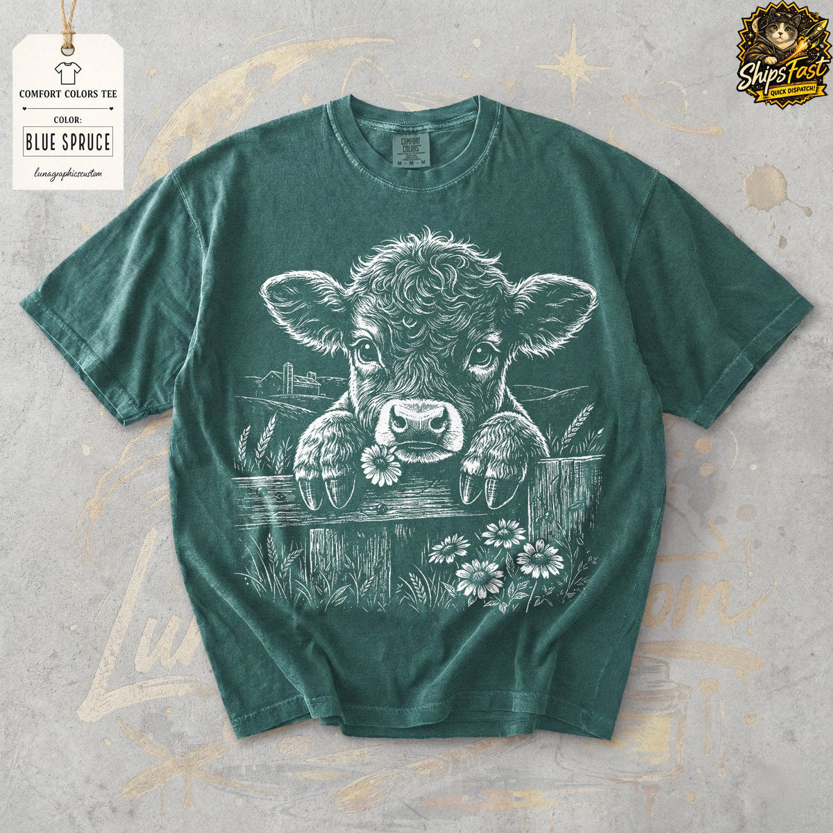 May include: Blue Spruce Comfort Colors tee featuring a white graphic of a calf peeking over a wooden fence, holding a daisy in its mouth. The design includes a rural landscape and additional daisies. The shirt has a crew neck and short sleeves.