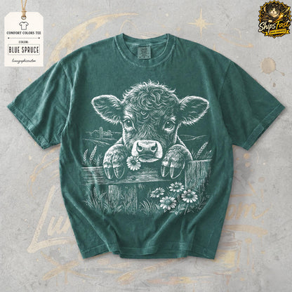 May include: Blue Spruce Comfort Colors tee featuring a white graphic of a calf peeking over a wooden fence, holding a daisy in its mouth. The design includes a rural landscape and additional daisies. The shirt has a crew neck and short sleeves.