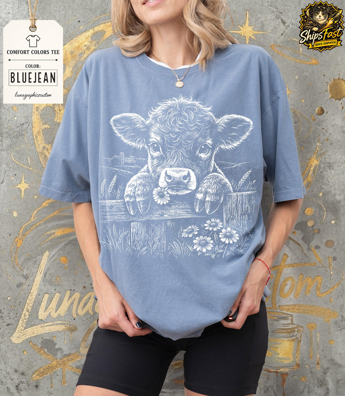 May include: Bluejean Comfort Colors tee featuring a white graphic of a cow peering over a fence with daisies. The shirt has short sleeves and a crew neck. The text "Comfort Colors Tee" and "Bluejean" are visible on a tag.