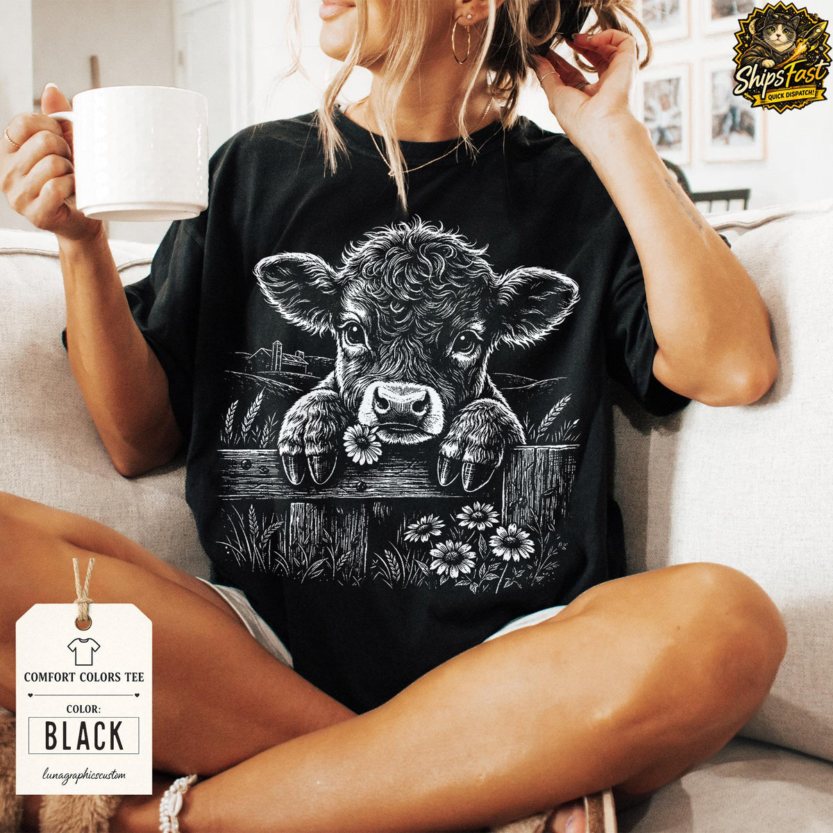 May include: Black Comfort Colors tee featuring a detailed graphic of a calf peering over a wooden fence, holding a daisy. The design includes wheat and daisies. The tag indicates the shirt is black.