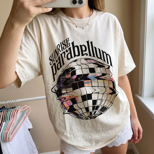 May include: A cream-colored short-sleeve t-shirt featuring a black graphic of a distorted disco ball. The text "SUNRISE parabellum" is printed in black above the disco ball graphic. The design includes colorful, glitch-like patterns within the ball.