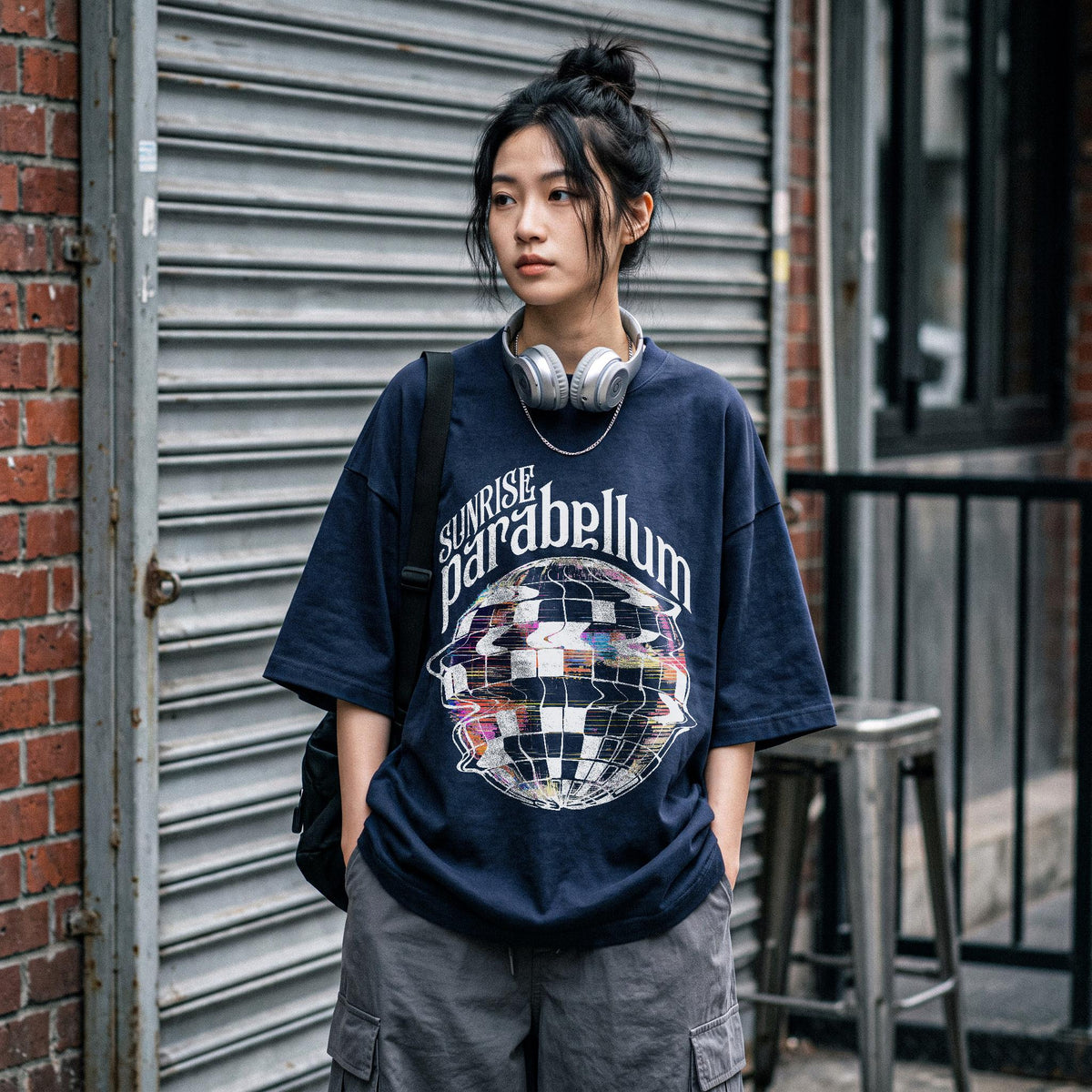 May include: A navy blue short-sleeve t-shirt featuring a graphic of a distorted, checkered sphere and the text "SUNRISE parabellum" in white lettering. The shirt has a loose, boxy fit and is paired with grey cargo pants and silver headphones.