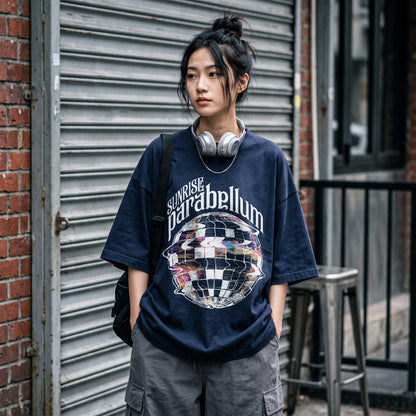 May include: A navy blue short-sleeve t-shirt featuring a graphic of a distorted, checkered sphere and the text "SUNRISE parabellum" in white lettering. The shirt has a loose, boxy fit and is paired with grey cargo pants and silver headphones.