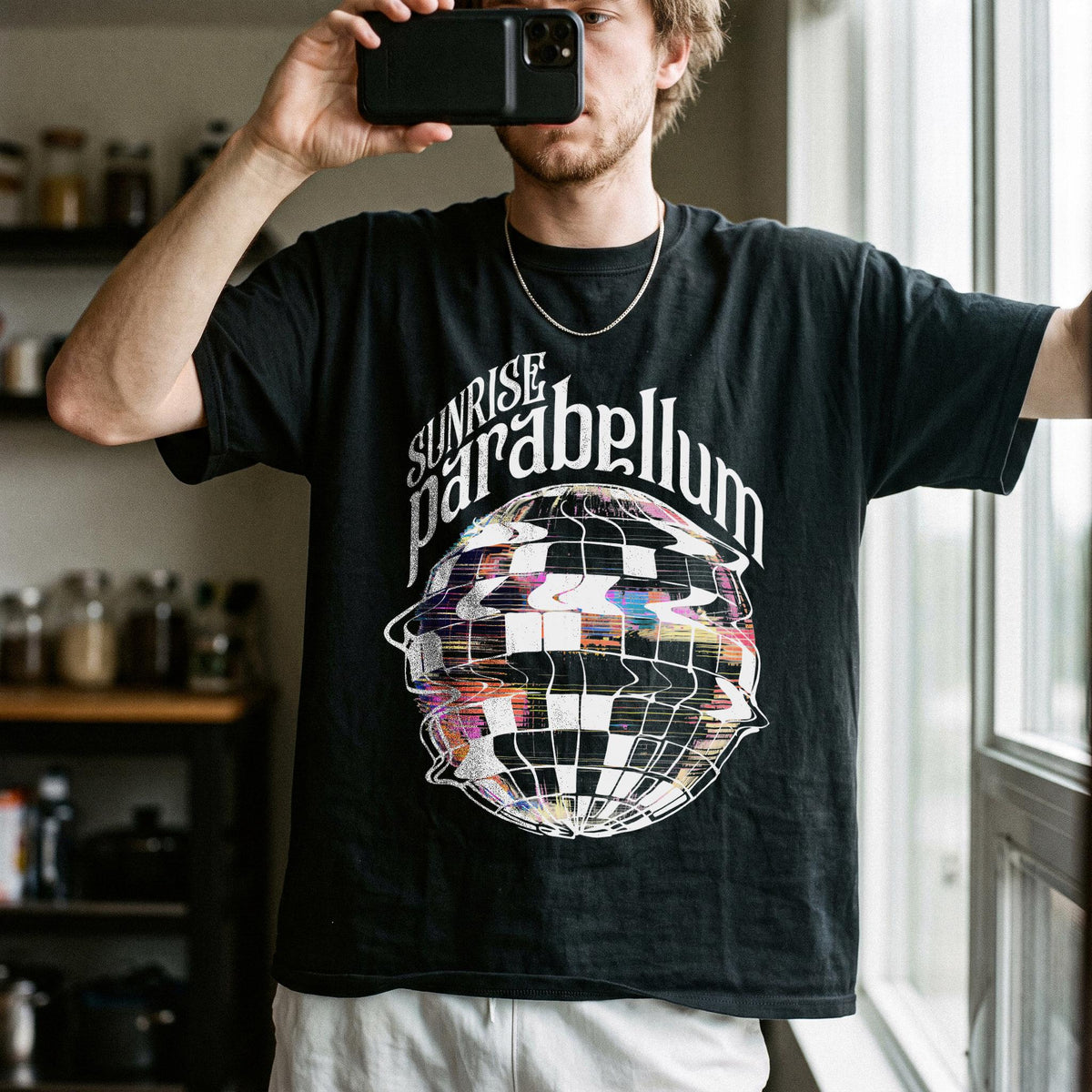May include: A black short-sleeve t-shirt featuring the white text "SUNRISE parabellum" arched over a graphic of a distorted, checkered disco ball with colorful, glitch-like pixelated accents throughout the sphere design.