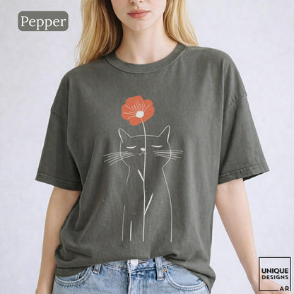 May include: A charcoal gray short-sleeve t-shirt featuring a white line art illustration of a cat with closed eyes. A single orange flower is positioned above the cat's head. The text Pepper is visible in the top left corner and Unique Designs Ari in the bottom right corner.