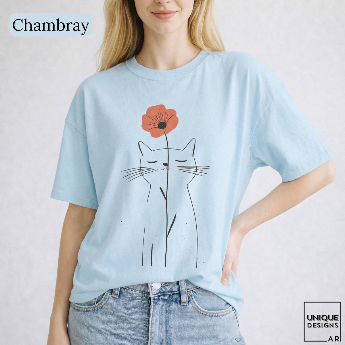 May include: A light blue t-shirt featuring a minimalist black line drawing of a cat with closed eyes. A single orange-red flower is positioned directly above the cat's head, with its stem extending downward in front of the cat's face.