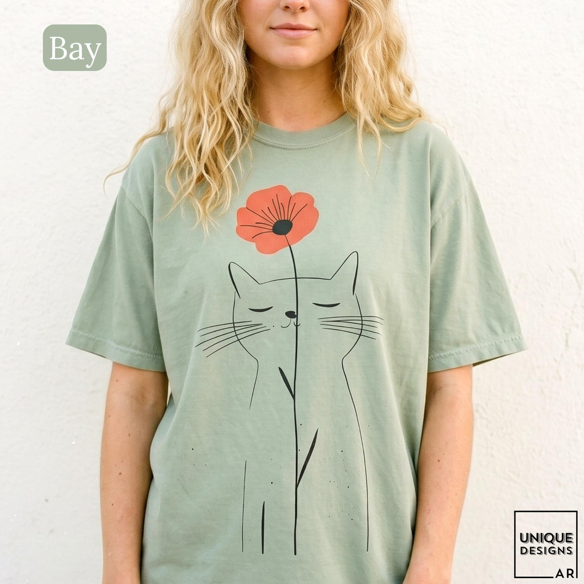 May include: A sage green short-sleeve t-shirt featuring a black line art illustration of a cat with closed eyes. A single orange poppy flower is positioned above the cat's head, with its stem extending downward along the cat's body.